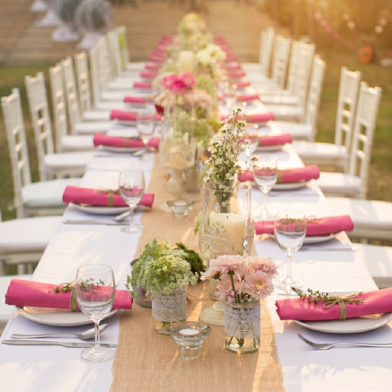 Long outdoor table set for a meal with white chairs, pink napkins, and floral centerpieces.